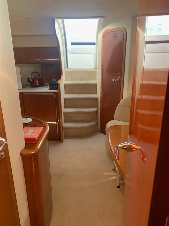 Interior view of a yacht showing a carpeted lounge area, stairs leading up, and kitchen space with a kettle.